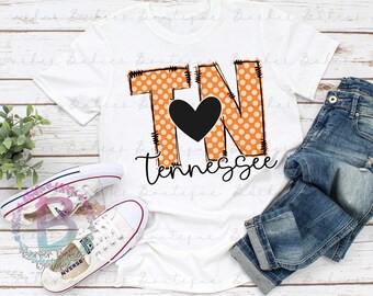 May include: White t-shirt with the letters "TN" in orange polka dots and a black heart. The word "Tennessee" is written below in a script font. The shirt is styled with blue jeans and white trainers.