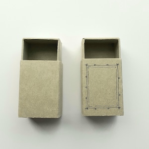 May include: Two open, rectangular matchboxes. One is plain, the other has a black, hand-drawn frame design. Both are made of a light brown, textured paper material. The boxes are positioned against a white background.