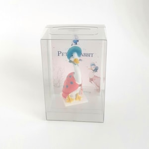 May include: A goose figurine in a clear display box. The goose wears a blue hat and a red cape with blue polka dots. The figurine has yellow feet and a white body. The box has the words "Peter Rabbit" printed on it.