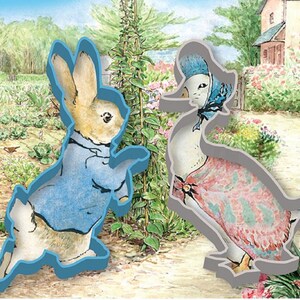 May include: Two cookie cutters, one shaped like a rabbit wearing a blue jacket and the other shaped like a duck wearing a pink dress. Both cutters are grey.