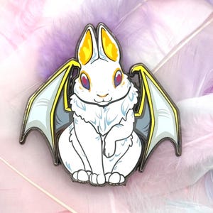 May include: White rabbit with yellow and gold wings, sitting on a pink and purple feather background. The rabbit has red eyes and a fluffy white body.