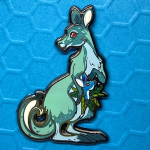 May include: A teal blue enamel pin of a kangaroo carrying a smaller kangaroo in its pouch. The larger kangaroo has a white tail and red eyes. The smaller kangaroo is blue and has a white tail. Both kangaroos are surrounded by green leaves.