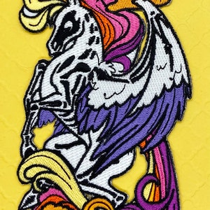 May include: Embroidered patch featuring a white skeletal winged unicorn with a rainbow mane and tail. The unicorn is standing on a cloud with a sun above it.