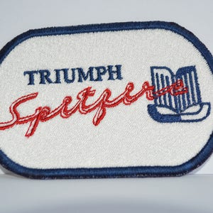 Könnte beinhalten: Ein weißer, ovaler gestickter Aufnäher mit blauem Rand. Der Aufnäher zeigt den Text "TRIUMPH" in Blau und "Spitfire" in Rot. Rechts auf dem Aufnäher befindet sich ein stilisiertes blaues Bild einer Kühlergrill.