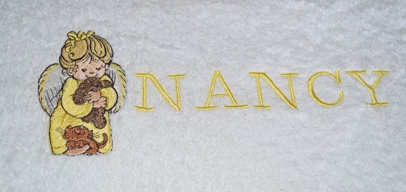 K&ouml;nnte beinhalten: Wei&szlig;es Handtuch mit einem gestickten Bild eines Engels, der einen Teddyb&auml;r und eine Katze h&auml;lt. Der Name "NANCY" ist unter dem Bild gestickt.