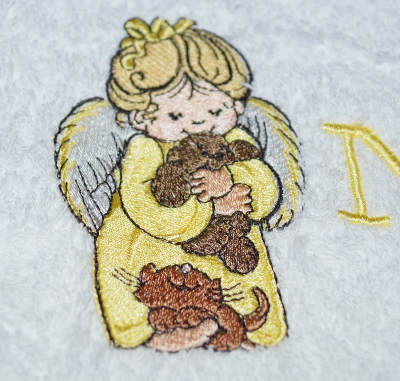 K&ouml;nnte beinhalten: Wei&szlig;es Handtuch mit Stickerei, auf dem ein gelber Engel einen braunen Teddyb&auml;r und eine braune Katze h&auml;lt. Der Engel hat blonde Haare und einen gelben Heiligenschein. Das Handtuch hat den Buchstaben "I" in Gelb in der oberen rechten Ecke gestickt.