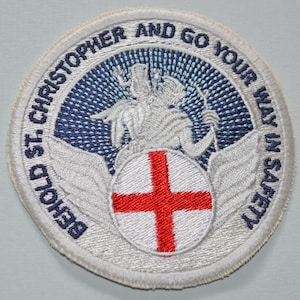 May include: White embroidered patch with a blue and white background. The patch features a white figure with wings and a red cross on a white circle. The text "Behold St Christopher and go your way in safety" is embroidered around the edge of the patch.