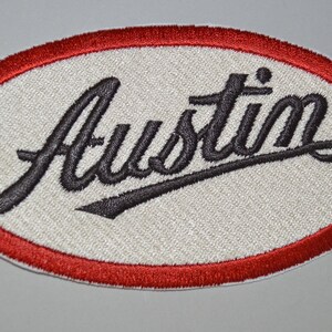 Könnte beinhalten: Ein weißer ovaler Patch mit rotem Rand. Das Wort "Austin" ist in schwarzer Kursivschrift gestickt.