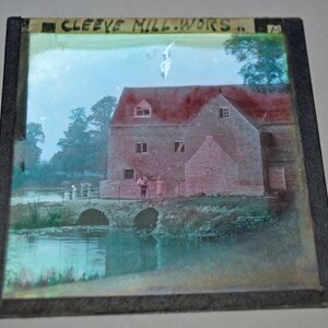 Op de afbeelding: Een vintage foto van een bakstenen gebouw met een rood dak, een brug en een rivier. Op het gebouw staat "CLEEVE MILL.WORS.". Er staan drie mensen voor het gebouw.