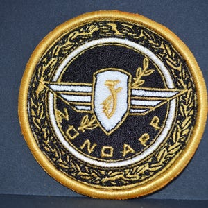 Op de afbeelding: Een gouden geborduurde patch met een zwarte achtergrond en witte letters. De patch heeft een wit schild met een gouden letter "J" erin, geflankeerd door witte vleugels met gouden versieringen. De woorden "Juno App" staan in een witte cirkelvormige rand rond het schild en de vleugels.