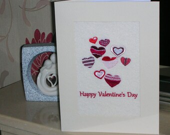 May include: A white card with a red felt insert. The insert has a cluster of pink, red, and white embroidered hearts. The text "Happy Valentine's Day" is embroidered in red at the bottom of the card.