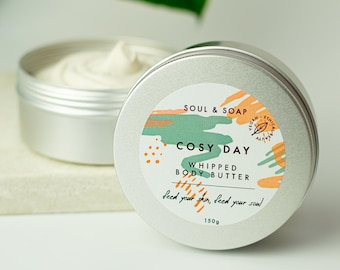 May include: A silver tin with a white label that reads "SOUL & SOAP COSY DAY WHIPPED BODY BUTTER feed your skin, feed your soul 150g". The label has a colorful abstract design with green, orange, and pink hues.