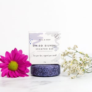 Op de afbeelding: Een paarse shampoo bar met de tekst "OH SO SILVER! SHAMPOO BAR" op de verpakking. De verpakking vermeldt ook "SOUL & SOAP" en "love your hair, respect your world". De bar ligt op een wit oppervlak met een roze bloem en witte bloemen.