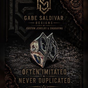 May include: A silver ring with a unique, angular design. The ring is set against a dark background with the text "Gabe Saldivar Designs" and "Often Imitated Never Duplicated". The ring has a complex, geometric shape with dark accents.