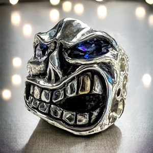 May include: A silver ring with a detailed skull design. The skull has a wide, toothy grin and two blue gemstones for eyes.