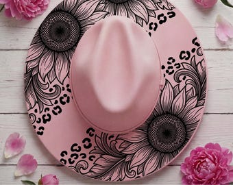 May include: A light pink fedora hat featuring a black line art design of large sunflowers and leopard print spots across the brim. The hat has a structured crown and a wide, flat brim, showcasing a detailed botanical and animal pattern.