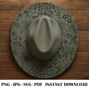 May include: An olive green fedora hat with a wide brim. The brim features a floral and leopard print design in black. The hat is set against a wooden background.