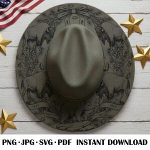 May include: A sage green fedora hat with a detailed black illustration of bison, a bear, and floral elements. The hat has a wide brim and a structured crown. Gold star decorations and an American flag are in the background.