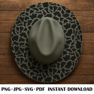 May include: A fedora hat with a wide brim, featuring a unique black and olive green pattern. The hat's crown is olive green, and the brim has a repeating pattern. The hat is displayed on a wooden surface.