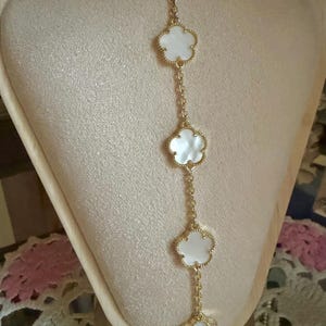 Op de afbeelding: Een goudkleurige ketting met vijf witte klavervormige hangers. Elke hanger heeft een goudkleurige rand en is verbonden door fijne gouden kettingen. De ketting wordt getoond op een beige sieraden buste.