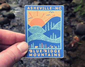 Op de afbeelding: Een rechthoekige sticker met een blauwe achtergrond met een gestileerd berglandschap met een opkomende zon. De tekst "Asheville NC" staat bovenaan en "Blue Ridge Mountains" onderaan. De sticker heeft een witte rand.