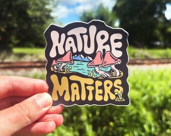 Könnte beinhalten: Ein Aufkleber mit schwarzem Hintergrund und buntem Text, der "Nature Matters" lautet. Der Text ist in einer verspielten Schriftart gehalten und von einer Szene mit einem See mit Bergen im Hintergrund und rosa Pilzen im Vordergrund umgeben.