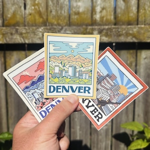 May include: Three colorful stickers featuring illustrations of cityscapes. The top sticker is yellow with a cityscape and the word "DENVER" in a blue box. The left sticker is pink with a cityscape and the word "DENVER" partially visible. The right sticker is red with a cityscape and the word "YER" partially visible.