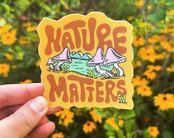Könnte beinhalten: Ein gelber Aufkleber mit dem Text "Nature Matters" in braunen Buchstaben. Der Aufkleber zeigt eine Cartoon-Illustration von Pilzen, die in der Nähe eines Gewässers wachsen.