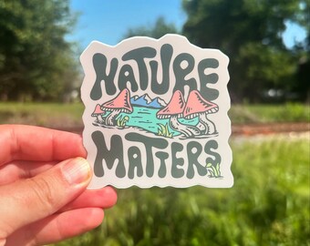 Könnte beinhalten: Ein Aufkleber mit grünem und weißem Hintergrund, auf dem der Text "Nature Matters" in einer verspielten Schriftart steht. Der Aufkleber zeigt außerdem eine Cartoon-Illustration von drei rosa Pilzen mit weißen Punkten, die in der Nähe eines blauen Sees mit grünen Bergen im Hintergrund wachsen.