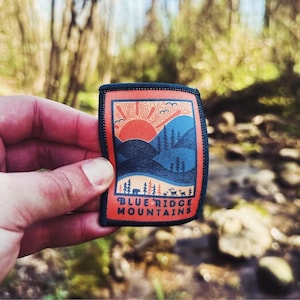 Puede incluir: Un parche negro y naranja con una imagen estilizada de una puesta de sol sobre montañas. El texto "BLUE RIDGE MOUNTAINS" está impreso debajo de la imagen.