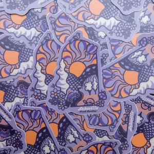 May include: A stack of purple and orange stickers featuring a stylized illustration of the state of North Carolina with the text "Asheville - North Carolina" in a white banner. The illustration features a sun, mountains, trees, and a car driving on a winding road.