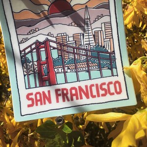 May include: A sticker with a cartoon illustration of the Golden Gate Bridge and the San Francisco skyline. The sticker is on a light blue background with the text "SAN FRANCISCO" in red.