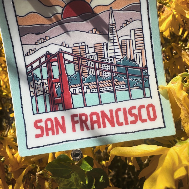 May include: A sticker with a cartoon illustration of the Golden Gate Bridge and the San Francisco skyline. The sticker is on a light blue background with the text "SAN FRANCISCO" in red.