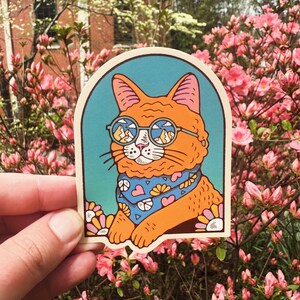 Pu&ograve; includere: Un adesivo con un gatto arancione dei cartoni animati che indossa occhiali rotondi con un paesaggio montano riflesso nelle lenti. Il gatto indossa un bandano blu con cuori rosa e un disegno di fiori gialli. Lo sfondo &egrave; un gradiente blu e verde.
