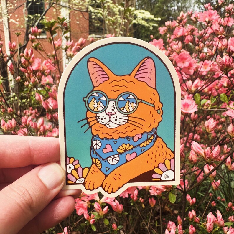 Pu&ograve; includere: Un adesivo con un gatto arancione dei cartoni animati che indossa occhiali rotondi con un paesaggio montano riflesso nelle lenti. Il gatto indossa un bandano blu con cuori rosa e un disegno di fiori gialli. Lo sfondo &egrave; un gradiente blu e verde.