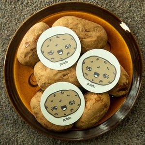 Puede incluir: Un bol de cerámica marrón lleno de patatas. Cada patata tiene una pegatina con una cara de patata de dibujos animados y la palabra "patata" impresa debajo.