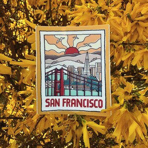 May include: A yellow sticker with a retro illustration of the Golden Gate Bridge and the San Francisco skyline. The illustration is set against a sunset with a red sun and the text "San Francisco" at the bottom.