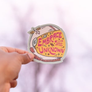 May include: A sticker featuring a cartoon astronaut helmet with the text "Embrace the Unknown" in pink and yellow lettering. The helmet is decorated with stars and planets.