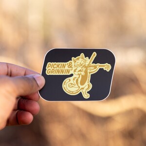 May include: A black and gold sticker with a cat playing a violin. The text on the sticker reads "Pickin' & Grinnin'".