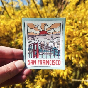 May include: A sticker with a retro illustration of the Golden Gate Bridge and the San Francisco skyline. The sticker has a blue background and the text "SAN FRANCISCO" in red.