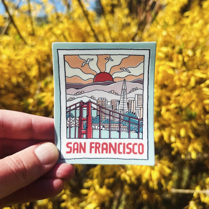 May include: A sticker with a retro illustration of the Golden Gate Bridge and the San Francisco skyline. The sticker has a blue background and the text "SAN FRANCISCO" in red.