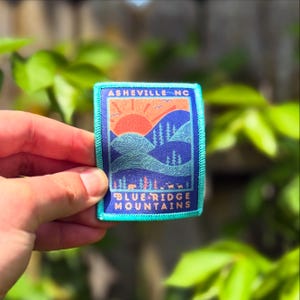 May include: A rectangular patch with a teal border featuring a mountain landscape design. The patch includes text that reads "Asheville NC" and "Blue Ridge Mountains". The design incorporates a rising sun, trees, and rolling hills in shades of orange, blue, and teal.