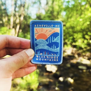 May include: Blue and orange embroidered patch with the text "Asheville, NC" and "Blue Ridge Mountains". The patch features a stylized mountain range with a sun rising over the peaks.