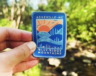 May include: Blue and orange embroidered patch with the text "Asheville, NC" and "Blue Ridge Mountains". The patch features a stylized mountain range with a sun rising over the peaks.