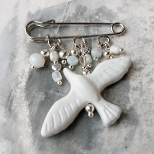 May include: A white ceramic dove brooch with a silver safety pin and dangling silver and white beads.