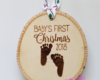 Może przedstawiać: Drewniana ozdoba choinkowa z napisem "Baby's First Christmas 2018" i dwoma śladami stóp dziecka. Ozdoba jest przymocowana do białej wstążki z czerwonymi i zielonymi lampkami.