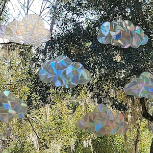 May include: Six cloud-shaped decorations made from iridescent materials hang in front of a tree. The clouds reflect a rainbow of colors, creating a shimmering effect. The decorations are likely for home decor.