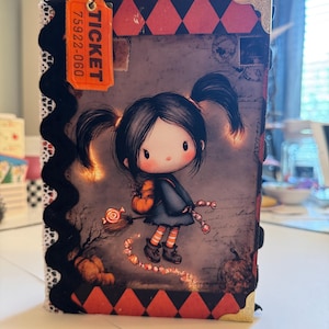 May include: A rectangular journal featuring a cover illustration of a cartoon character with pigtails holding a broomstick and candy. The cover has a black and red diamond border, white lace trim, silver corner protectors, and an orange ticket tag.