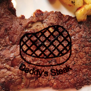 Può includere: Primo piano di una bistecca alla griglia con un motivo a griglia bianco e nero e il testo "Daddy's Steak" sulla superficie.