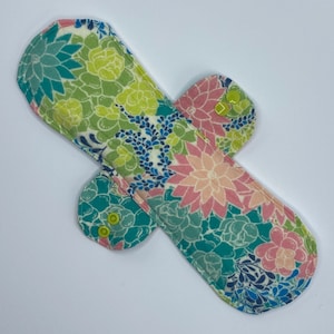May include: A reusable cloth menstrual pad with a floral print in shades of green, pink, and blue. The pad has two wings with snaps for attaching to underwear.